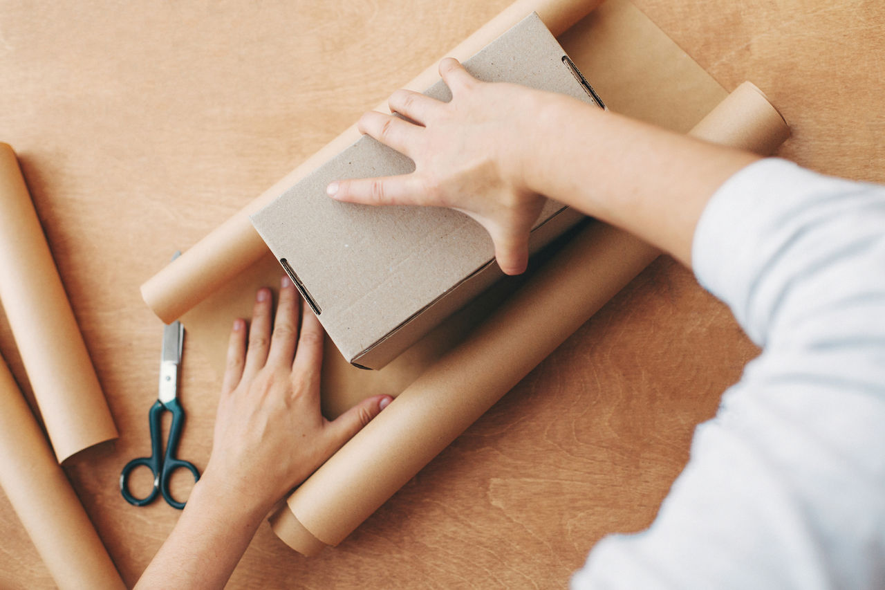 Wrapping christmas gifts concept. Hands wrapping christmas gift in craft paper and pine branches, cones, gingerbread cookies, thread, cinnamon, scissors on rural wooden table