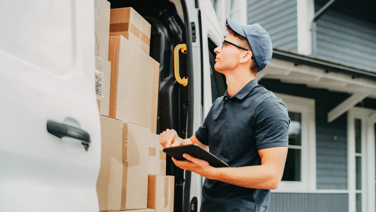 Courier Using Tablet Computer Next to Open Delivery Van Side Door with Cardboard Parcels. Mailman Delivering the Package to a Homeowner.