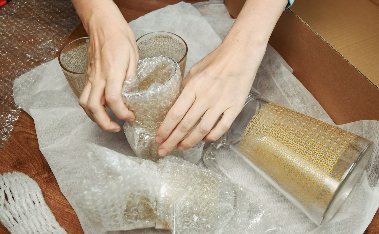 Woman hands pack up breakable fragile glassware and wrap into wrapping bubble plastic in preparation to move to new home. Packing up jug and drink glassses